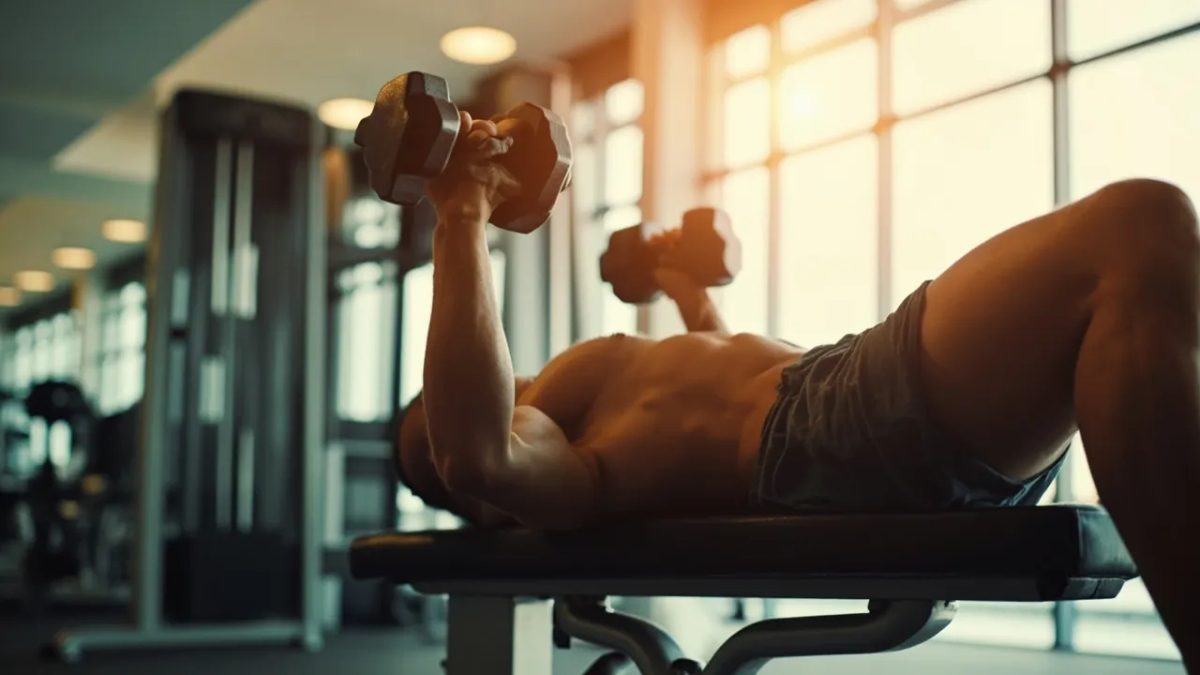 Hombre realizando press de banca con mancuernas en un gimnasio luminoso, ejercicio de pectorales.