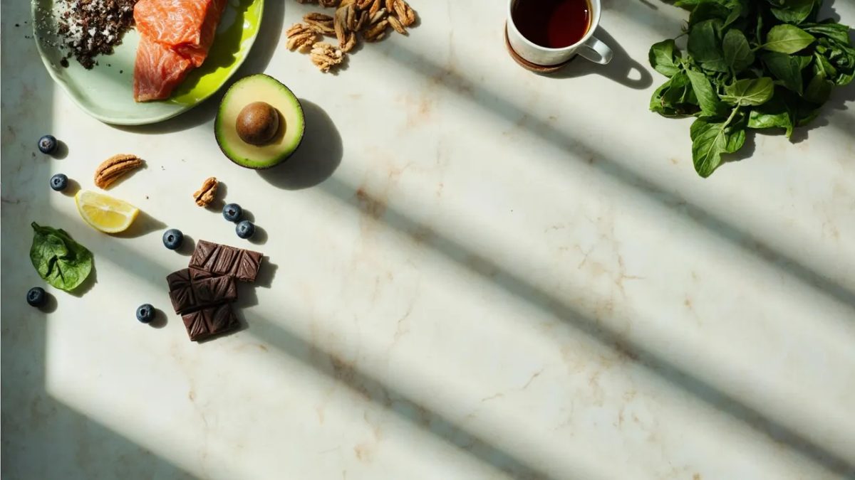 Disposición cenital de ingredientes saludables sobre una superficie clara: medio aguacate, un trozo de salmón, frutos secos, arándanos, una taza de té verde, chocolate negro, rodajas de limón y espinacas frescas.