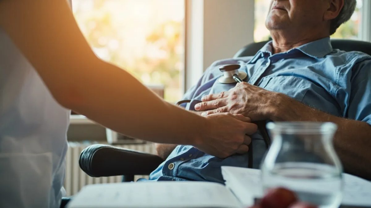 Médico auscultando el corazón de un paciente adulto sentado en una consulta, con el estetoscopio apoyado sobre el pecho y luz natural cálida en el ambiente.