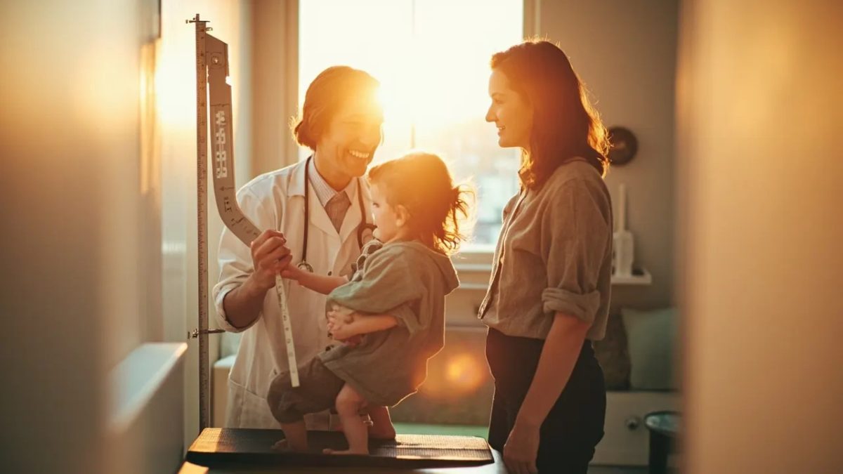 Pediatra diversa midiendo la estatura de una niña en una consulta luminosa, con su madre sonriendo a su lado.