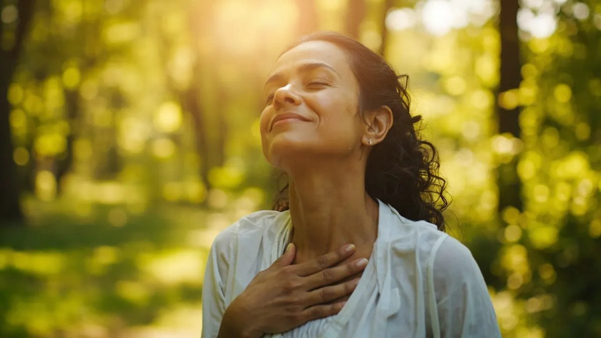 Persona diversa al aire libre tomando una respiración profunda con la mano apoyada suavemente sobre el pecho, rodeada de vegetación y luz cálida del atardecer