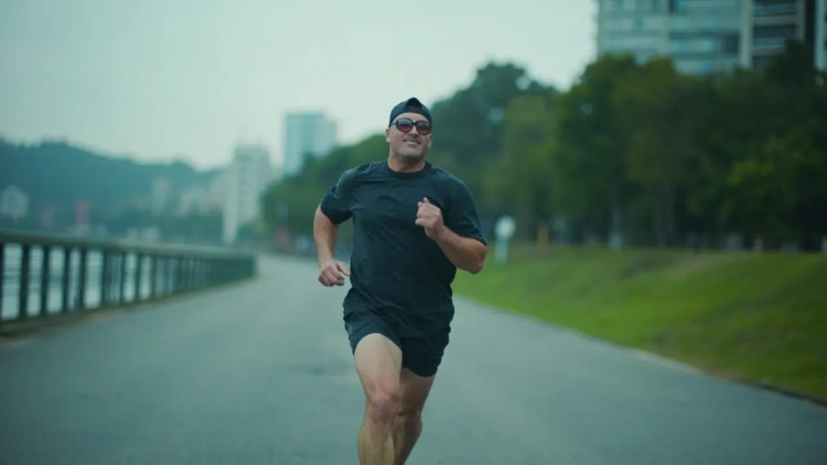 Hombre de unos cuarenta años trotando por un sendero urbano junto a un río a primera hora de la mañana, bajo una luz fría y difusa de día nublado, vestido con ropa deportiva oscura, con árboles y edificios modernos desenfocados al fondo.